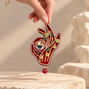 Peut inclure: Une broche en forme de main en velours rouge profond, ornée d'un œil brodé avec un iris bleu au centre. La surface est décorée de petites perles florales jaunes et bleu sarcelle, d'accents dorés et d'une perle rouge unique suspendue.