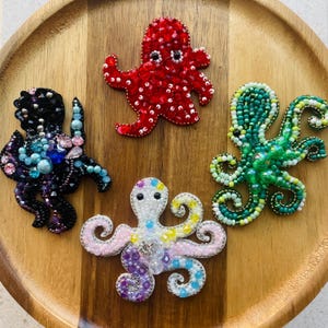 Puede incluir: Cinco broches de pulpo con cuentas de varios colores se muestran en una bandeja de madera. Los pulpos son rojos, negros, verdes, blancos y una mezcla de rosa, morado y amarillo. Cada broche tiene un diseño único y está hecho con pequeñas cuentas.