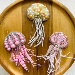 Puede incluir: Tres broches de medusa hechos a mano con intrincados abalorios. Un broche muestra perlas blancas y amarillas, otro presenta cuentas rosas con detalles plateados, y el tercero incluye perlas rosas suaves y plumas delicadas.