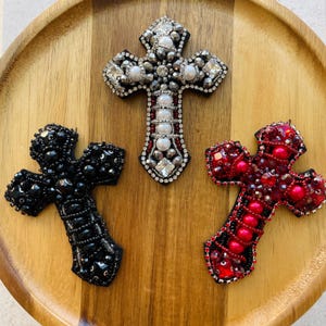 May include: Three beaded cross brooches in black, silver, and red, each with intricate detailing. The crosses are embellished with beads and rhinestones, resting on a wooden tray. The silver cross features pearls and clear crystals.
