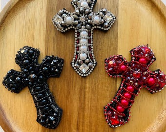 May include: Three beaded cross brooches in black, silver, and red, each with intricate detailing. The crosses are embellished with beads and rhinestones, resting on a wooden tray. The silver cross features pearls and clear crystals.