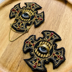 May include: Two black cross-shaped embroidered patches featuring gold thread scrollwork, red crystal accents, and a central realistic blue eye with a gold eyelid. One patch includes a thin gold chain draped across the bottom edge.