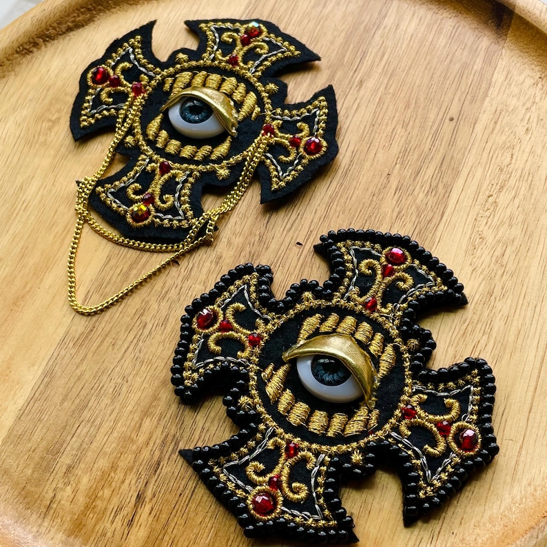 May include: Two black cross-shaped embroidered patches featuring gold thread scrollwork, red crystal accents, and a central realistic blue eye with a gold eyelid. One patch includes a thin gold chain draped across the bottom edge.