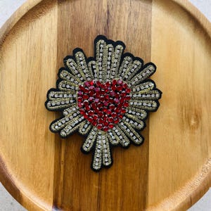 May include: A heart-shaped, embroidered patch with a black base. The heart is filled with red rhinestones, surrounded by gold and silver beaded accents. The patch is on a wooden surface.