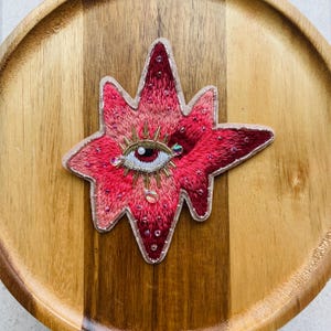 May include: Embroidered star-shaped patch with an eye design at its centre. The patch is in shades of red and pink, with gold and clear bead accents. The eye has a detailed iris and is surrounded by gold thread.