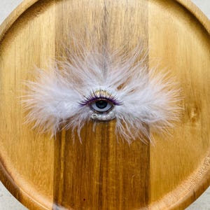 以下が含まれることがあります： 中央に暗い虹彩を持つリアルな目が配置された装飾的なブローチ。白い羽、紫色の先端を持つまつげ、小さな白いビーズの列、そして目の下部に配置された単一の透明なラインストーンのアクセントで構成されています。