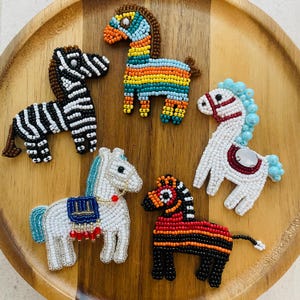 May include: A collection of beaded horse and zebra figurines in various colors. The figurines are arranged on a wooden tray. The horses are white, blue, red, and orange. The zebra is black and white.