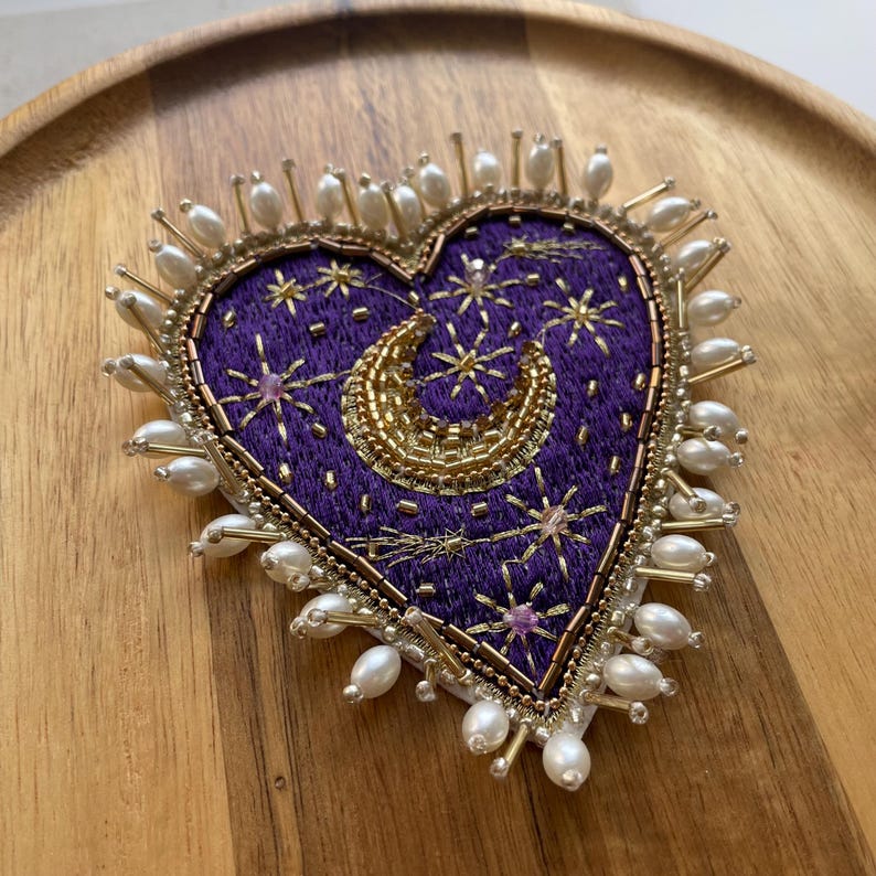 Pu&ograve; includere: Spilla ricamata a forma di cuore con sfondo viola scuro. Il design presenta una luna crescente dorata e stelle, con perline dorate e perlate che delineano il cuore. I bordi sono adornati con perline simili a perle.