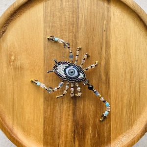 May include: A beaded brooch shaped like a spider with a central evil eye motif. The body features a blue and white eye surrounded by dark beads, with multiple radiating legs crafted from clear, metallic, and light blue glass beads and crystals.