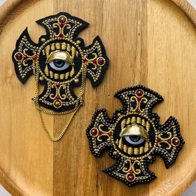May include: Two black cross-shaped patches featuring intricate gold embroidery, red bead accents, and a central realistic eye with a gold eyelid. One patch includes a gold-toned metal chain draped across the front, adding texture and dimension.