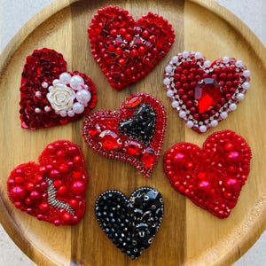 May include: A collection of handmade heart-shaped brooches in various shades of red and black. Some hearts are embellished with beads, sequins, and crystals. One heart features a white rose and pearl accents. The brooches are displayed on a wooden tray.