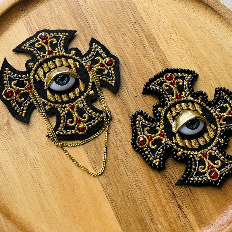May include: Two black felt cross-shaped patches featuring gold embroidery, red rhinestones, and a central realistic eye with a gold eyelid. One patch includes a gold-toned metal chain draped across the front, while the other is plain.
