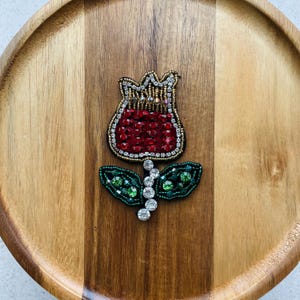 Könnte beinhalten: Eine handgefertigte Blumenbrosche mit einem rot paillettenbesetzten Tulpendesign mit Gold- und Silberakzenten. Die Brosche enthält grüne, mit Perlen besetzte Blätter und klare Kristallakzente. Die Brosche liegt auf einer Holzoberfläche.