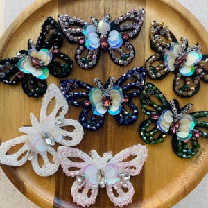 Könnte beinhalten: Eine Sammlung von aufgenähten Schmetterlingen mit Perlen in verschiedenen Farben, darunter Schwarz, Weiß, Rosa, Blau und Grün. Jeder Schmetterling hat schillernde Pailletten und glitzernde Kristalle, angeordnet auf einem Holztablett. Geeignet für Bastelarbeiten und Verzierungen.