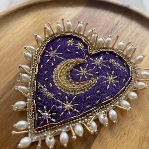 Pu&ograve; includere: Spilla ricamata a forma di cuore con un tema celeste. Il tessuto viola &egrave; adornato da stelle dorate, una luna crescente e perline dorate. I bordi sono rifiniti con perle e perline dorate.