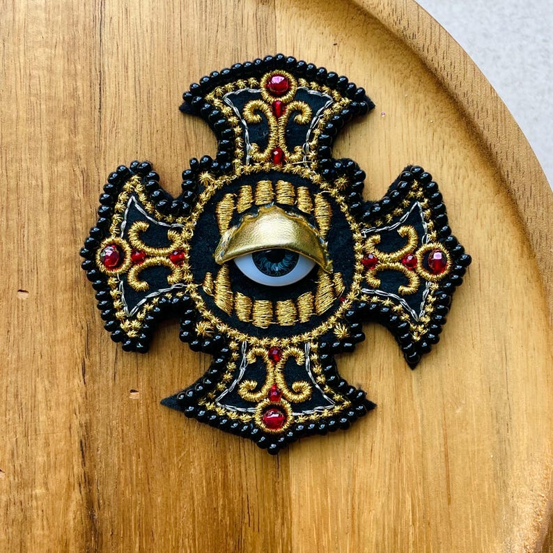 May include: A cross-shaped embroidered patch featuring black fabric with gold thread scrollwork, small black beads along the edges, and red circular accents. A central realistic blue eye with a gold eyelid is positioned in the middle of the cross.