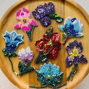 Op de afbeelding: Een collectie van acht handgemaakte kralen bloembroches in diverse kleuren, waaronder roze, paars, blauw, rood en wit. Elke broche is voorzien van ingewikkeld kralenwerk, pailletten en kristallen accenten in bloemvormen met stelen en bladeren.