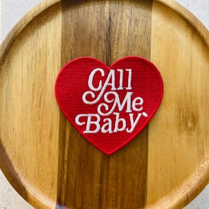 Könnte beinhalten: Ein roter herzförmiger Aufnäher mit den Worten "Call Me Baby" in weißer Schrift gestickt. Der Aufnäher ist auf einem Holztablett mit einem hellen und dunklen Holzmaserungsmuster zentriert.