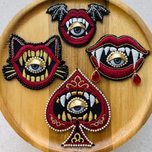 May include: Four embroidered patches featuring red lips, white fangs, and central realistic eyes. One patch is shaped like a spade, one like a cat face, one has bat wings, and one includes two red teardrop-shaped beads hanging from the lower lip.