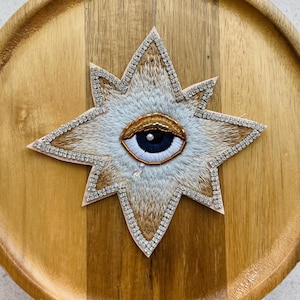 Peut inclure: Un écusson brodé en forme d'étoile présentant un motif d'œil central avec un iris bleu foncé, une sclère blanche et une paupière perlée dorée. L'étoile est bordée de strass argentés avec des détails en fil beige et blanc et une larme en cristal.