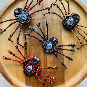 May include: Three spider-shaped beaded ornaments featuring central cabochon eyes. Each spider has a body crafted from dark beads and eight radiating legs made of wire strung with red, blue, amber, and black glass beads in various patterns.