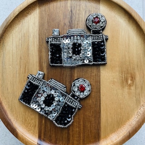 Puede incluir: Dos parches decorativos de cámara. Cada parche es negro con lentejuelas y cuentas plateadas. Una joya roja está centrada en el flash. Los parches están sobre una bandeja de madera.