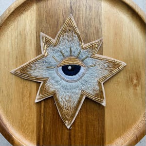 Op de afbeelding: Een stervormige geborduurde patch met een centraal oogmotief met een donkerblauwe pupil en een witte iris. Het ontwerp is geaccentueerd met metallic gouden kraaltjes langs de randen en stralen, met lichtbruin en wit stiksel door het geheel.