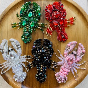 Puede incluir: Cinco broches de langosta con cuentas en verde, rojo, blanco, negro y rosa. Cada broche presenta un intrincado trabajo de cuentas, con detalles como pinzas, antenas y segmentos corporales. Los broches se exhiben en una bandeja de madera, mostrando la variedad de colores y diseños.