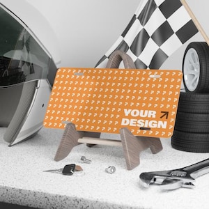 May include: An orange license plate with the text "YOUR DESIGN" and a right-pointing arrow, displayed on a wooden stand. A checkered flag, tires, and tools are in the background, suggesting an automotive theme.