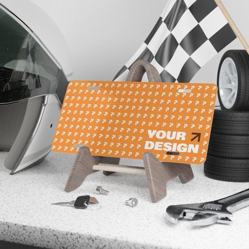 May include: An orange license plate with the text "YOUR DESIGN" and a right-pointing arrow, displayed on a wooden stand. A checkered flag, tires, and tools are in the background, suggesting an automotive theme.