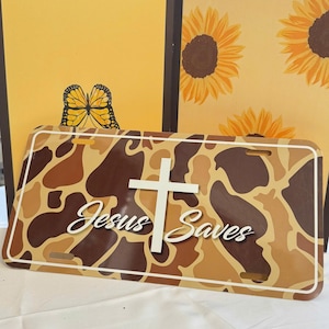 May include: License plate with a camouflage pattern in shades of brown and tan. The words "Jesus Saves" are written in white script, with a white cross above the text. The background includes yellow panels with butterfly and sunflower designs.