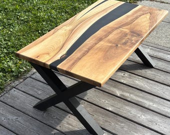 Op de afbeelding: Een rechthoekige houten salontafel met een zwarte epoxyharsinleg. Het tafelblad wordt ondersteund door zwarte, X-vormige metalen poten. De houtnerf is zichtbaar en de tafel is ongeveer 122 cm lang.