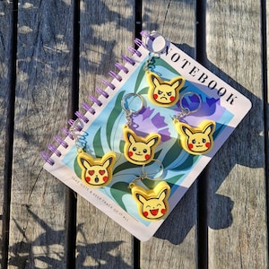 Puede incluir: Llaveros amarillos con temática de Pikachu con varias expresiones faciales, sujetos a llaveros plateados. Los llaveros están colocados sobre un cuaderno con la palabra "NOTEBOOK" visible. El cuaderno tiene una encuadernación en espiral morada y descansa sobre una superficie de madera.