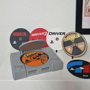May include: A gray PlayStation 1 console with a disc featuring the Crash Bandicoot logo. Several game discs are displayed, including Resident Evil, Driver 2, Driver, Duke Nukem, and a blue and red disc. The discs have the PlayStation logo.