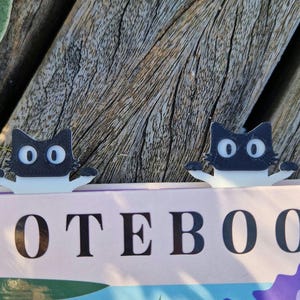 Könnte beinhalten: Zwei schwarze katzenförmige Lesezeichen mit weißen Bäuchen und großen, ovalen Augen sitzen auf einem Notizbuch. Das Wort "NOTEBOOK" ist in großen, schwarzen Buchstaben auf dem Einband des Notizbuchs aufgedruckt. Der Hintergrund ist eine verwitterte Holzoberfläche.
