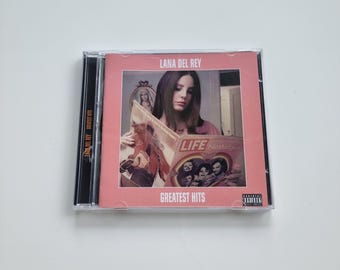 Puede incluir: Una caja de CD del álbum "Greatest Hits" de Lana Del Rey. La portada es rosa y muestra a Lana Del Rey leyendo una revista. La portada de la revista dice "LIFE Nostalgia". La caja del CD está en una funda de plástico transparente.