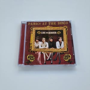Puede incluir: Una caja de CD con la banda Panic! At The Disco. La portada del álbum tiene un fondo granate con detalles dorados y el texto "Panic! At The Disco" en la parte superior. La imagen en el centro dice "Live in Denver" y presenta una foto de la banda.
