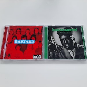 Può includere: Due custodie per CD affiancate. La custodia di sinistra ha una copertina rossa con la parola "BASTARD" in blu. La custodia di destra ha un'immagine in bianco e nero di una persona con la parola "CHROMATICA" in verde.