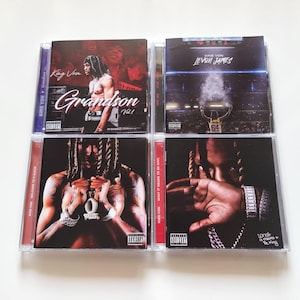 May include: Four King Von music CDs in clear plastic jewel cases arranged in a square grid. The covers feature photographic artwork of the artist, including titles Grandson Vol. 1, Levon James, Welcome to O'Block, and What It Means to Be King.