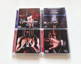 Puede incluir: Cuatro CD de música de King Von en cajas de plástico transparente dispuestas en una cuadrícula cuadrada. Las portadas presentan ilustraciones fotográficas del artista, incluidos los títulos Grandson Vol. 1, Levon James, Welcome to O'Block y What It Means to Be King.