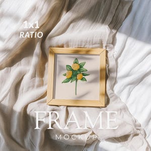 May include: A square light wood frame containing a print of a plant with three yellow flowers and green leaves. The text 1x1 RATIO is in the top left, and the text FRAME MOCKUP is centered at the bottom. The frame rests on a beige fabric.