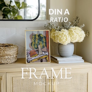 以下が含まれることがあります： 金色のフレームに入ったアートプリント。黄色い花、グラス、本のある静物画が描かれています。プリントは、織り込まれた前面を持つ明るい木製のキャビネットにあります。「FRAME MOCKUP」というテキストが下にあります。