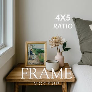 Peut inclure: Une œuvre encadrée dans un cadre doré, un vase rose avec une fleur et une tasse en céramique sont posés sur une table en bois. Un oreiller et un lit sont en arrière-plan. Le texte "FRAME MOCKUP" est en bas.