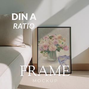 May include: A framed art print mockup featuring a floral still life in a glass vase. The artwork displays pink and white flowers with green foliage. The frame is black, and the text "DIN A RATIO FRAME MOCKUP" is overlaid on the image.