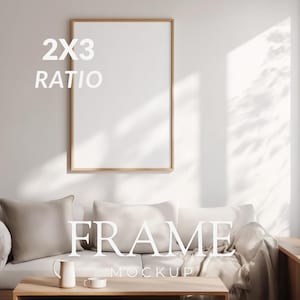 Op de afbeelding: Een houten frame 2x3 ratio blanco kunstafdruk mockup. Het frame hangt aan een witte muur boven een witte bank met kussens. De woorden "FRAME" en "MOCKUP" worden in witte tekst weergegeven.