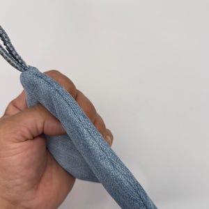 Può includere: Una pochette in tessuto denim azzurro di forma arrotondata con un passante in denim abbinato attaccato all'angolo superiore. L'articolo presenta cuciture visibili lungo il bordo curvo, che mostrano la trama testurizzata del materiale in cotone blu.