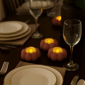 May include: A table setting with plates, silverware, and wine glasses. Three decorative, flower-shaped candles with a warm glow are placed on the table. A bouquet of flowers adds a touch of elegance to the scene.