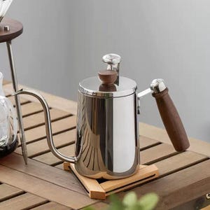 May include: A polished stainless steel gooseneck kettle with a wooden handle and lid knob. The kettle rests on a wooden trivet. The long, curved spout is a key feature. The design is modern and minimalist, ideal for pour-over coffee.