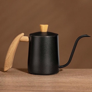 May include: A black gooseneck kettle with a wooden handle and lid knob. The kettle has a long, curved spout for precise pouring. The handle and lid knob are made of light-colored wood, contrasting with the black body. The kettle sits on a wooden surface.