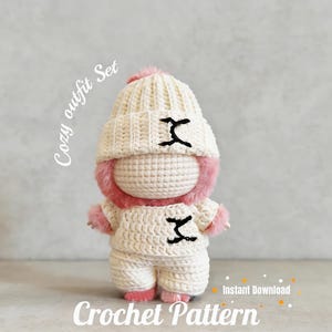 May include: A crochet doll wearing a cream-colored beanie with a pink pom-pom, a matching cream sweater, and cream pants. The sweater and beanie feature black stitched details. The doll has pink fuzzy trim around the face and hands. Text reads Cozy outfit Set and Crochet Pattern.